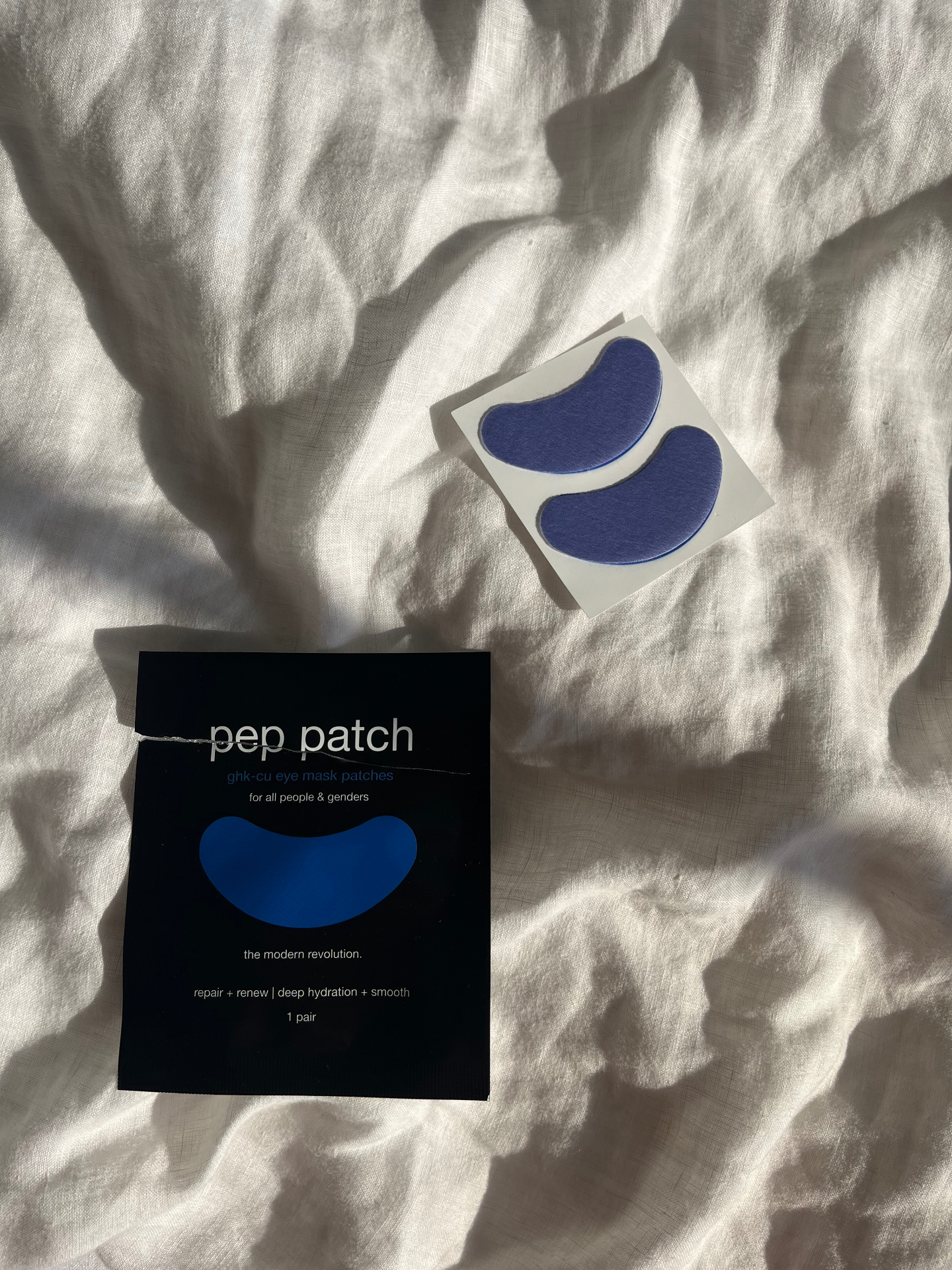 Pep Patch eye patches and black sachet resting on white bedding in morning sunlight